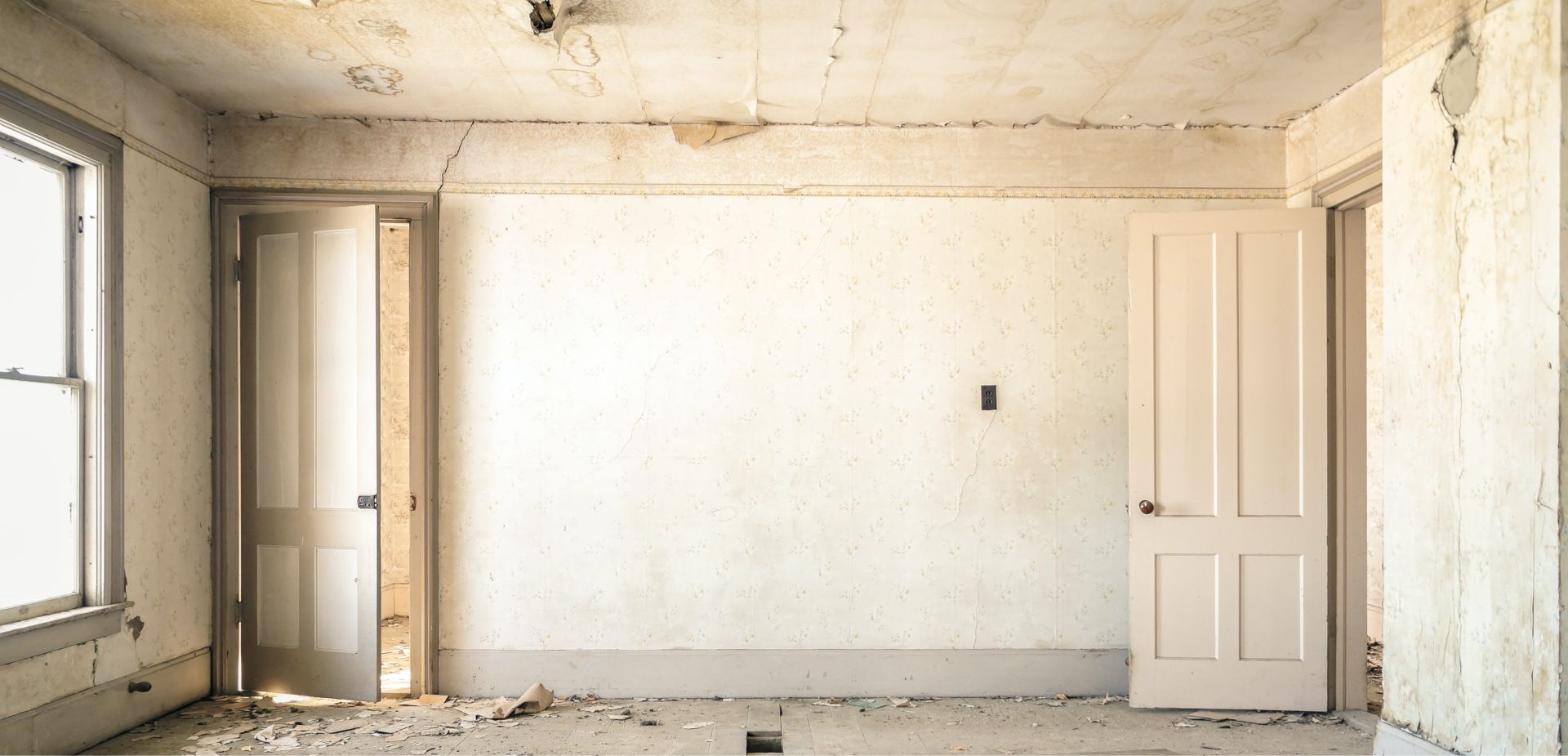Interior view of an abandoned room with two doors and a window, white walls, and a debris-covered floor.