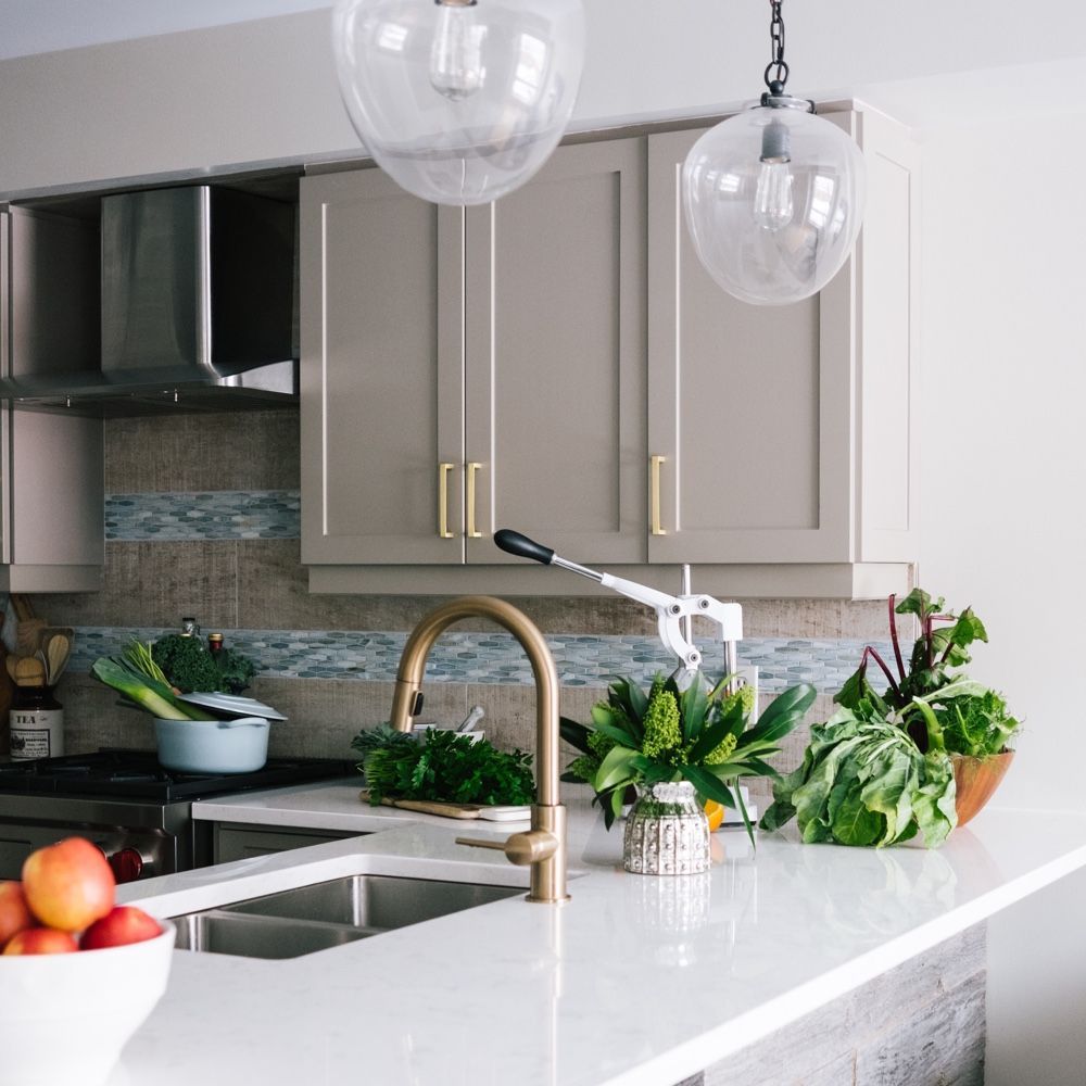 Modern kitchen with white countertop, brass faucet, fresh produce, and tan cabinets.