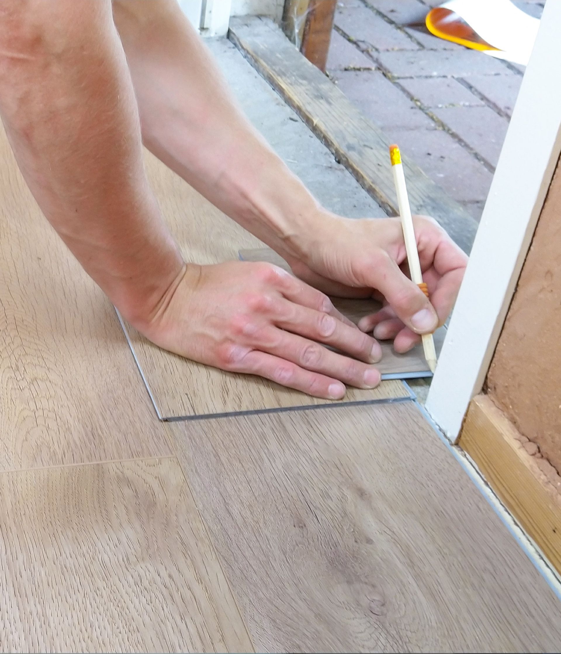Person using a pencil to trace a shape onto a flooring plank during installation, indoors.