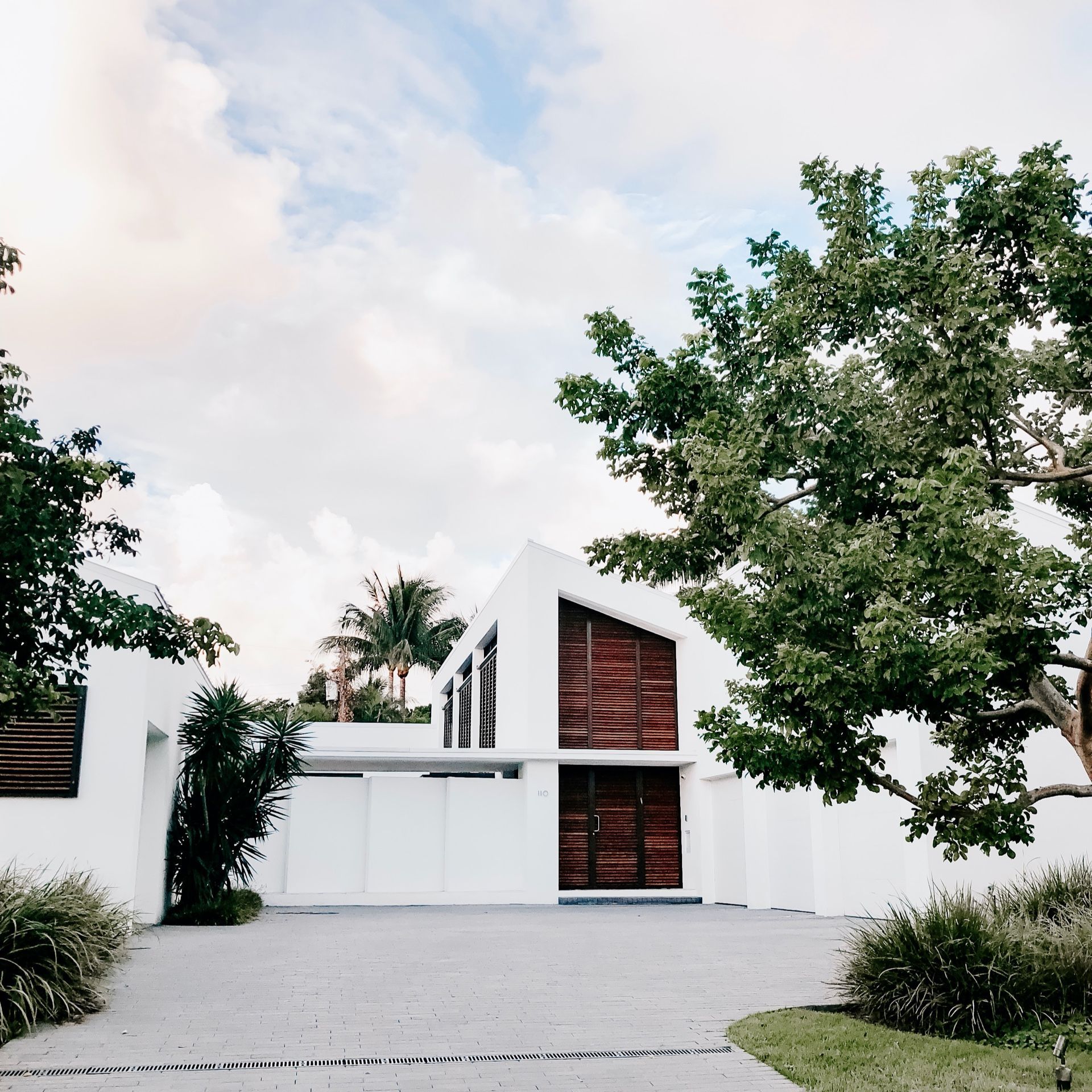 White modern house with wooden door and shutters, driveway, surrounded by trees under a cloudy sky.