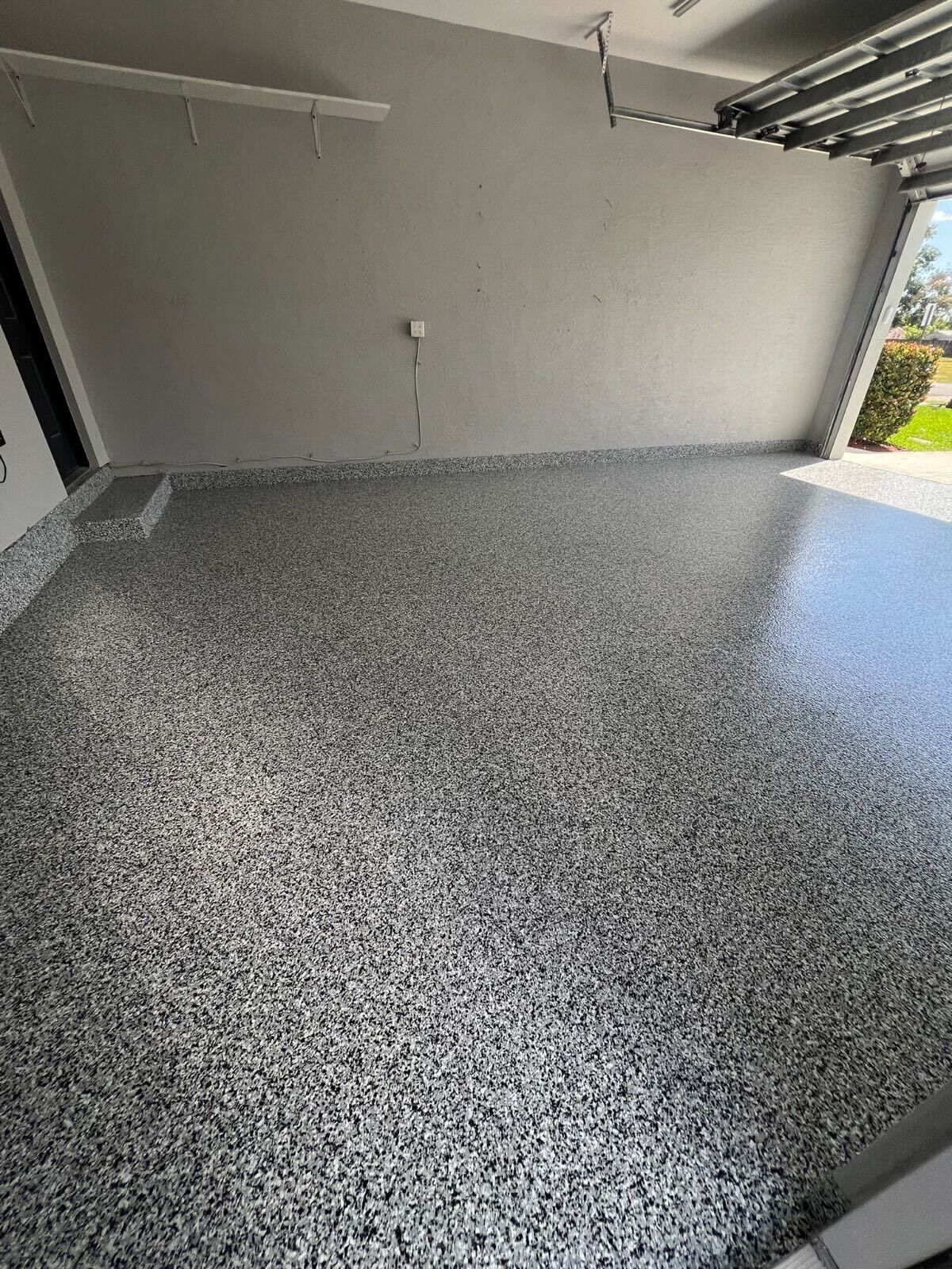 Empty speckled garage floor under an open garage door, showing a gray wall and daylight outside