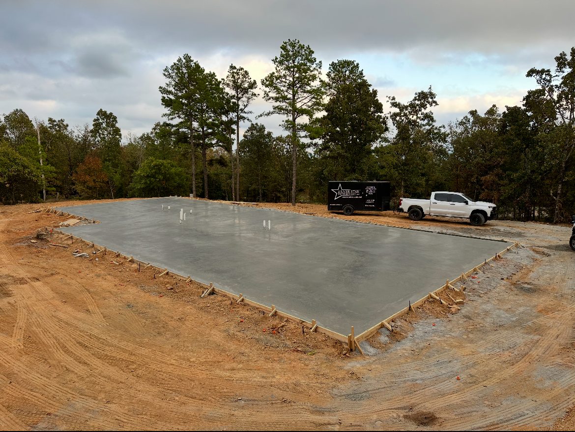 Concrete slab foundation at a wooded construction site with a parked truck and trailer nearby