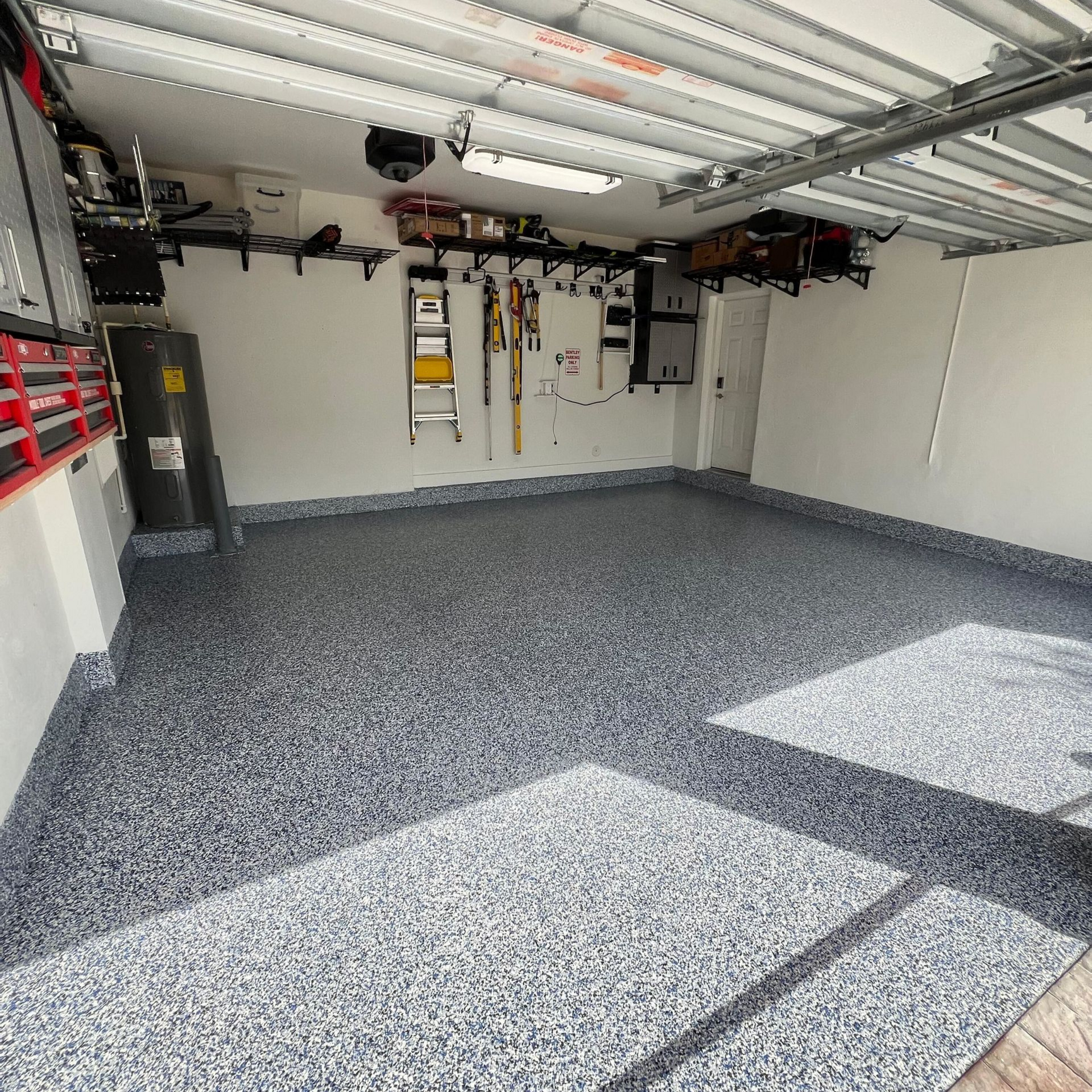 Empty garage with speckled gray floor, open door, overhead storage, and tools on the wall.