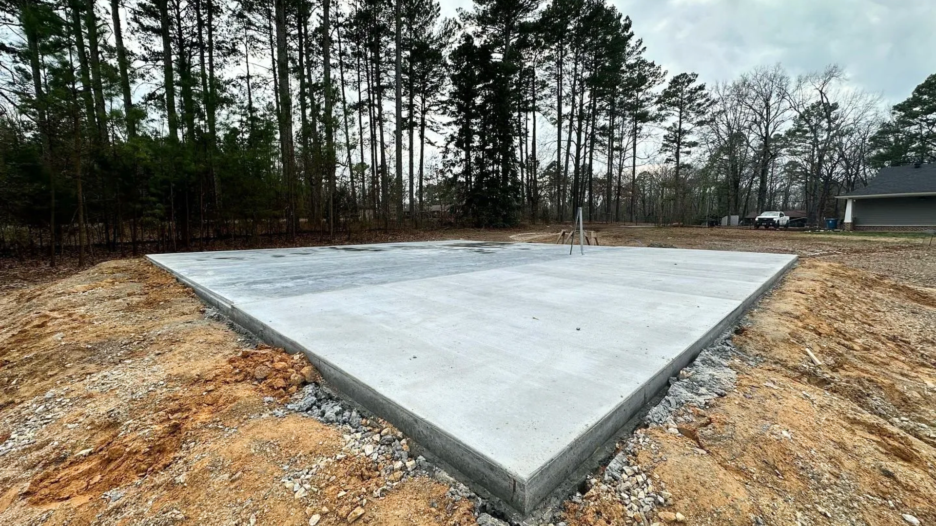 Concrete slab foundation on a cleared dirt lot beside a wooded tree line