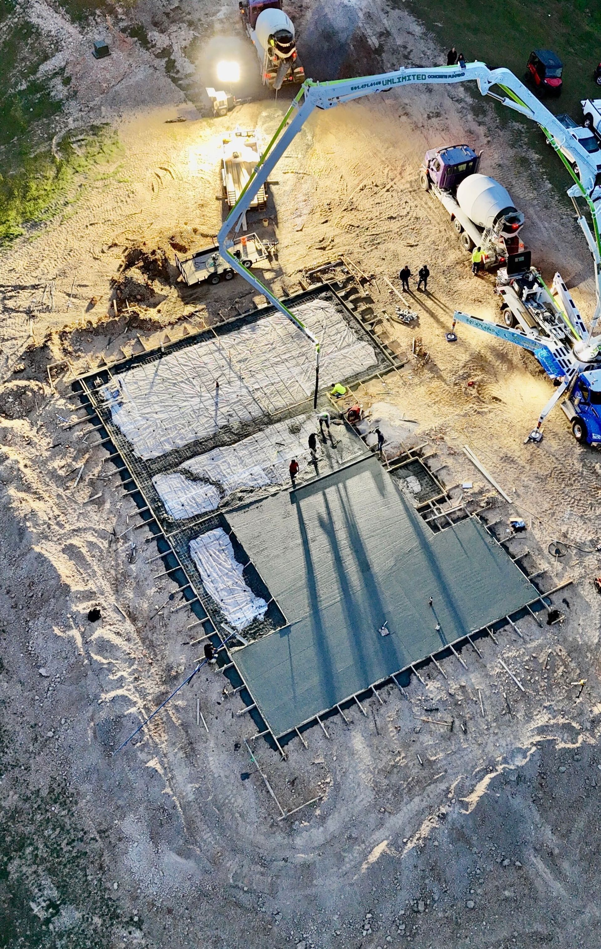Aerial view of a construction site with workers, a blue excavator arm, and a concrete foundation area.