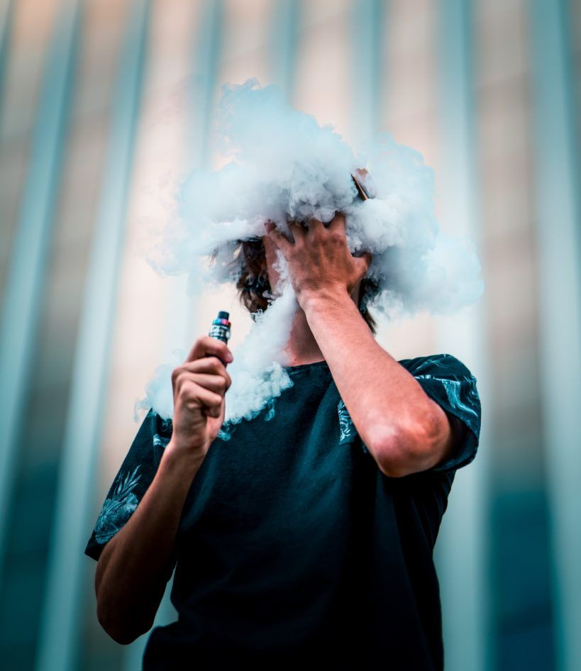 Person exhaling thick vapor cloud, covering face, holding vape pen. Blue-tinted background.