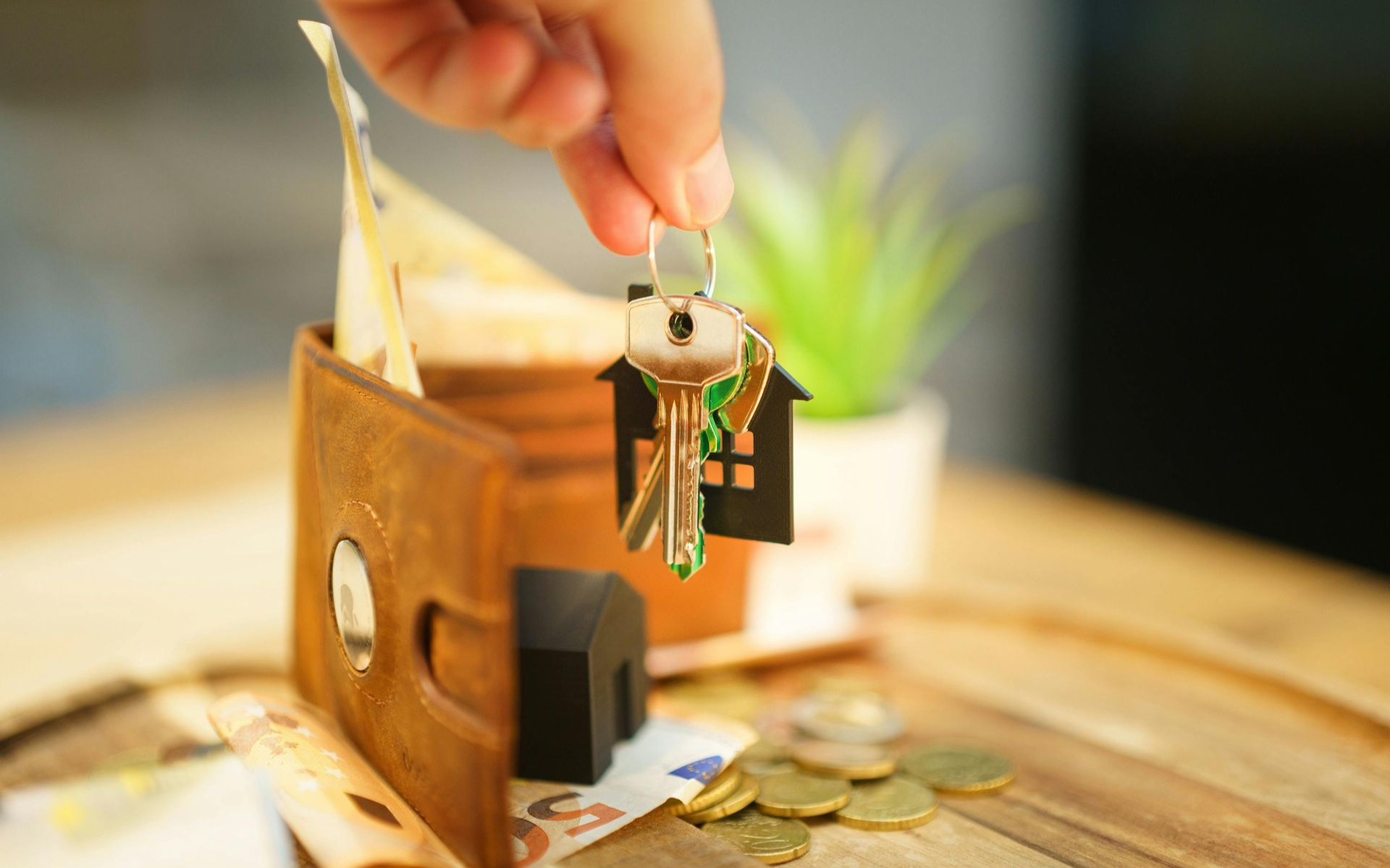 Hand holding keys with house charm, wallet with cash, and coins on wood table.