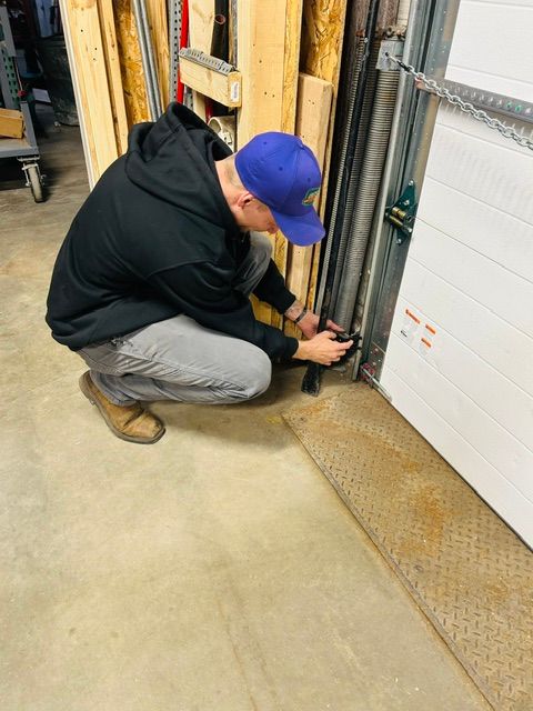 A person in a blue shirt repairs a garage door mechanism, attaching a cable to a bracket near the metal track.
