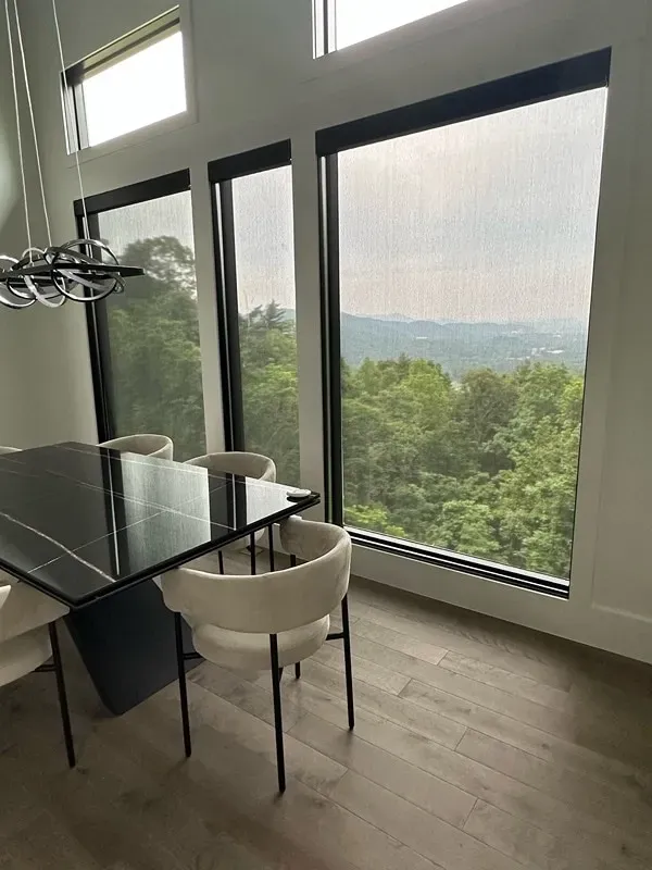 Modern dining area with a black table and white chairs overlooking a scenic forest and mountain view through large windows.