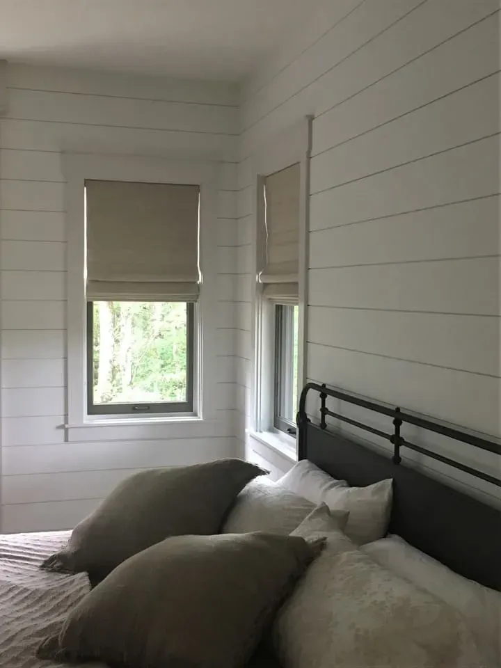 A bedroom with white shiplap walls, a dark metal bed frame, neutral-toned pillows, and two windows with tan roller shades.