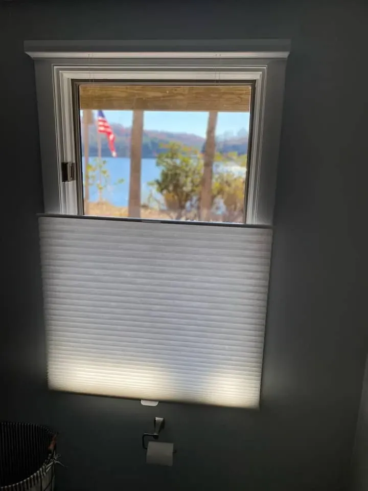 A window featuring a partially raised pleated white shade, revealing a view of a lake and an American flag outside.