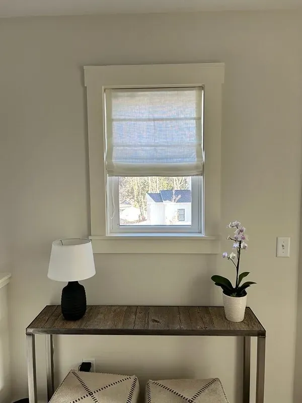 A lamp and an orchid sit on a narrow wooden console table in front of a white-framed window with a textured Roman shade.