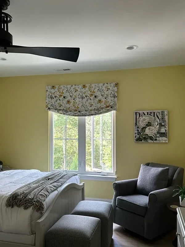 A cozy bedroom with pale yellow walls, a bed, two grey ottomans, a matching armchair, and a floral patterned window shade.