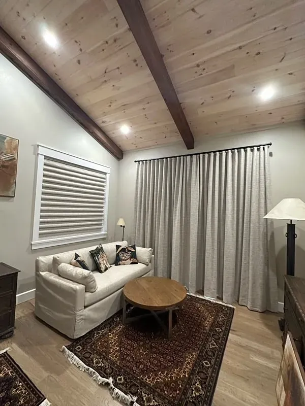 A living room with light wood floors, a beige sofa, patterned rug, wooden coffee table, large windows, and vaulted ceiling.