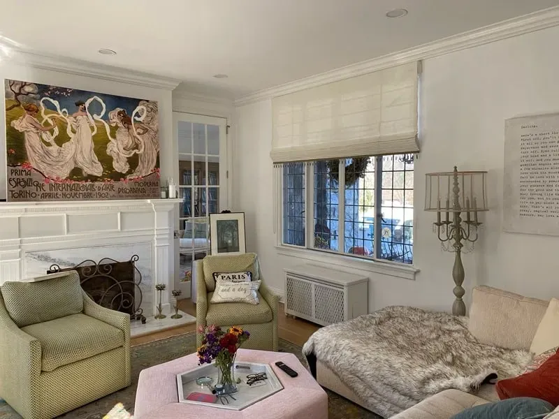 A cozy living room with a fireplace, large abstract artwork, a light green armchair, a pink ottoman, and a plush sofa.