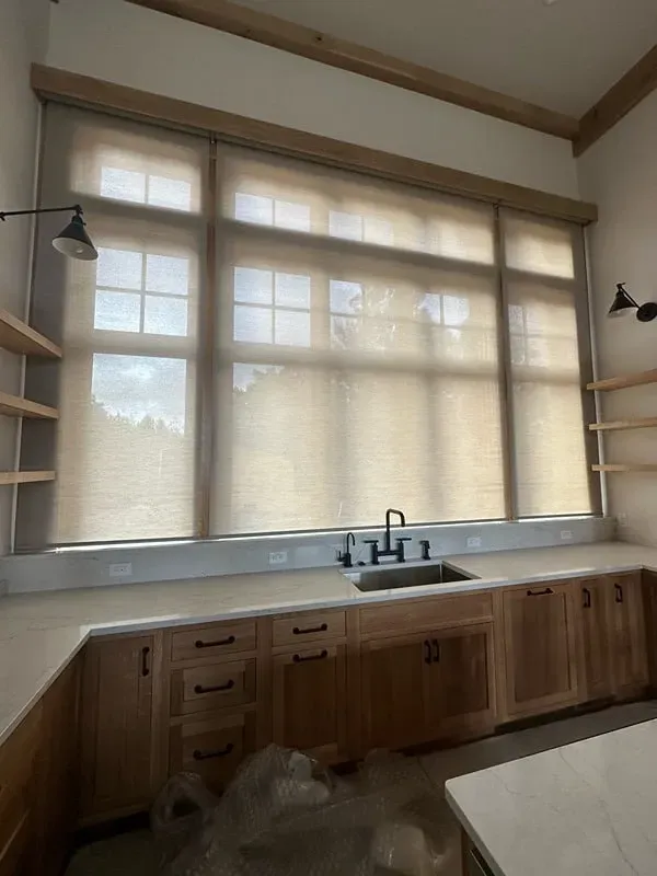 A modern kitchen sink area with light wood cabinets, open shelving, and large windows covered by beige roller shades.