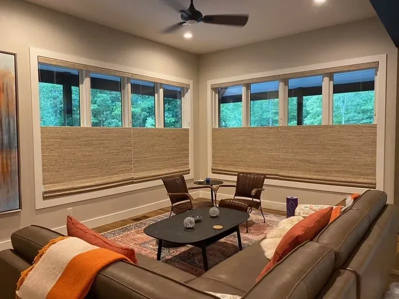 A living room with a large gray sectional, two chairs, a coffee table, and windows with woven shades overlooking trees.