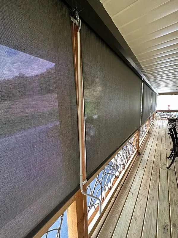 Dark grey mesh outdoor shades installed on a wooden deck, viewed from an angle showing the metal operation crank.