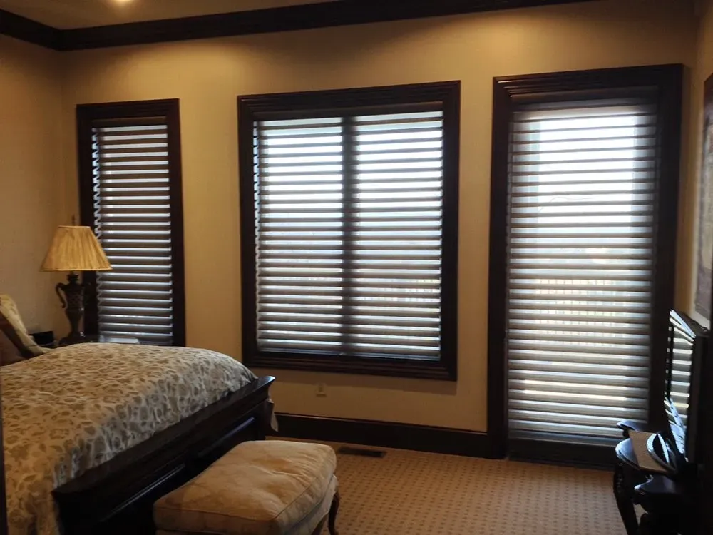 A bedroom featuring three windows with horizontal blinds, a dark wood bed frame, a patterned comforter, and a lamp.