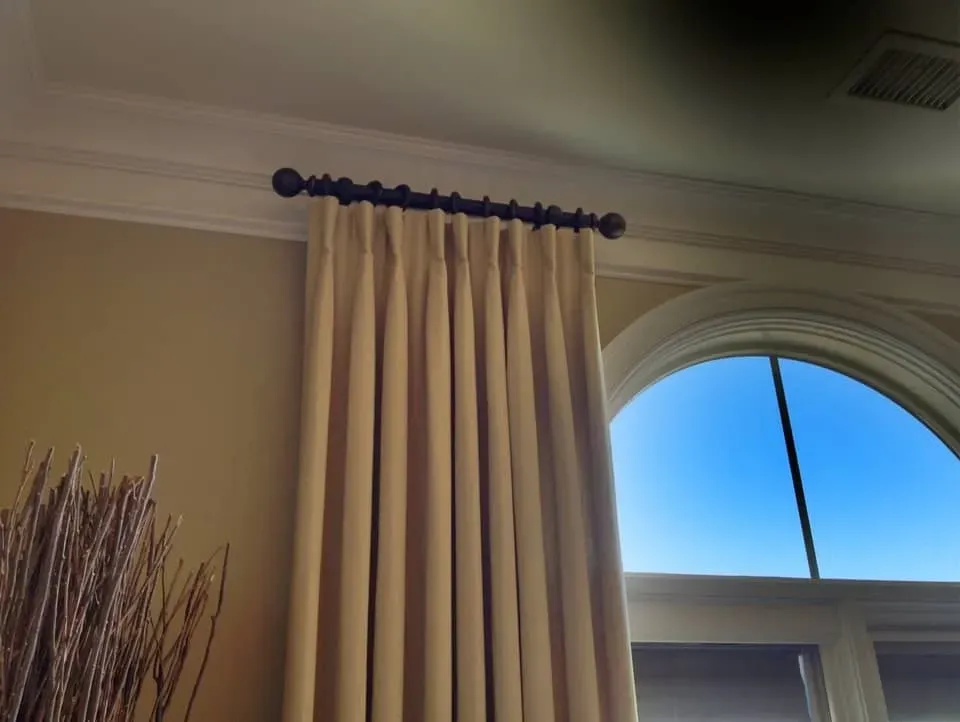 Beige pleated curtains hang from a dark decorative rod mounted near the ceiling next to an arched window.