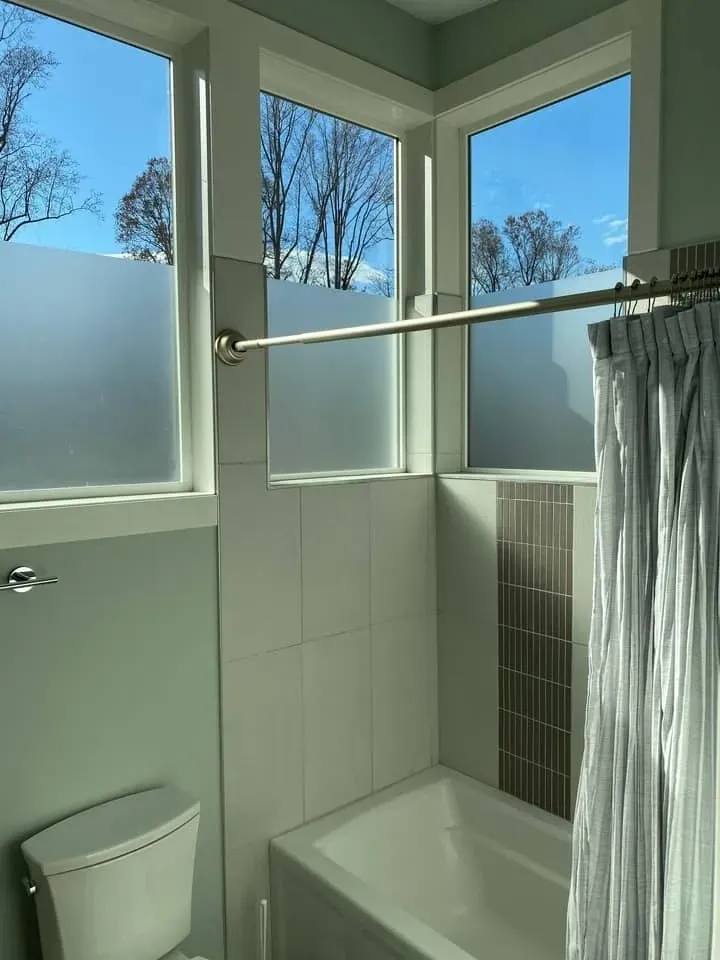 A corner bathroom featuring a white tub, a toilet, light walls, and windows with frosted glass and clear top panels.