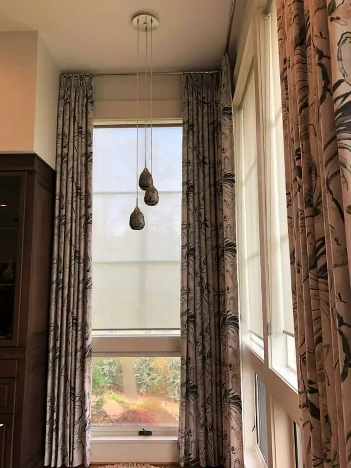 Floor-to-ceiling patterned curtains frame a tall window with shades, featuring a three-pendant light fixture above.