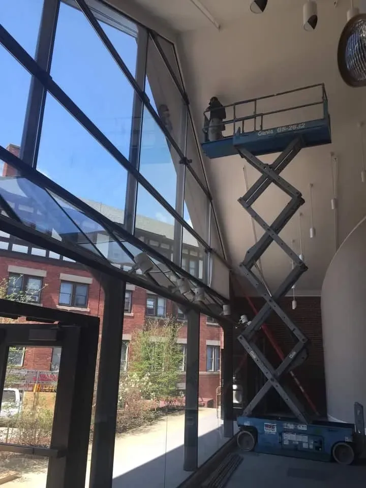 A worker stands on a blue scissor lift inside a building, reaching up to clean the large glass wall of the atrium.