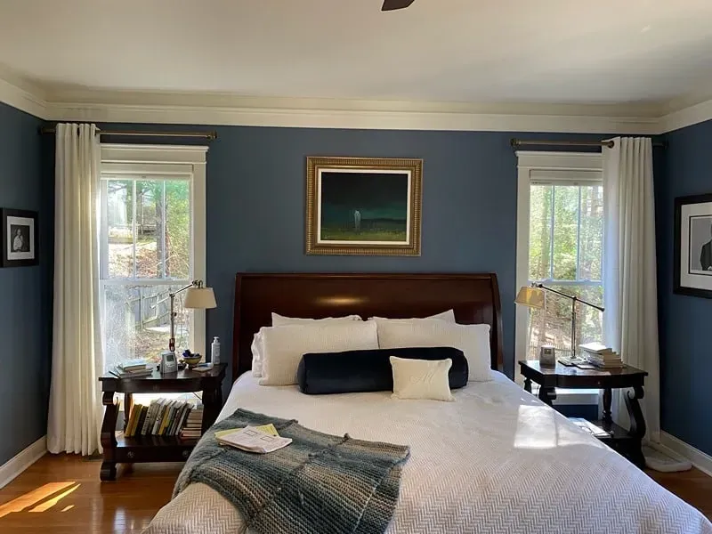 A bedroom with dark blue walls, white curtains, a wooden headboard, a white bedspread, and framed art on the walls.