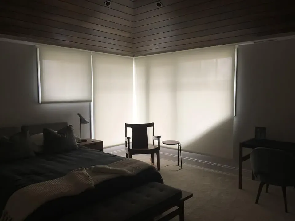 A modern bedroom with dark wood ceiling panels, a bed with dark linens, an accent chair, and large windows with blinds.