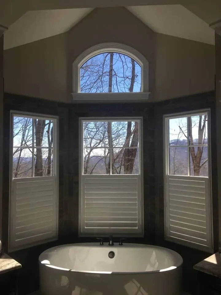 A white freestanding bathtub sits in front of three windows covered with white shutters beneath an arched transom window.