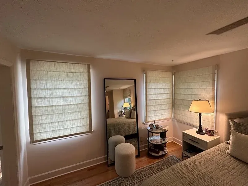 A bedroom corner with two tan textured roman shades, a full-length mirror, a small round shelf, and a nightstand with a lamp.