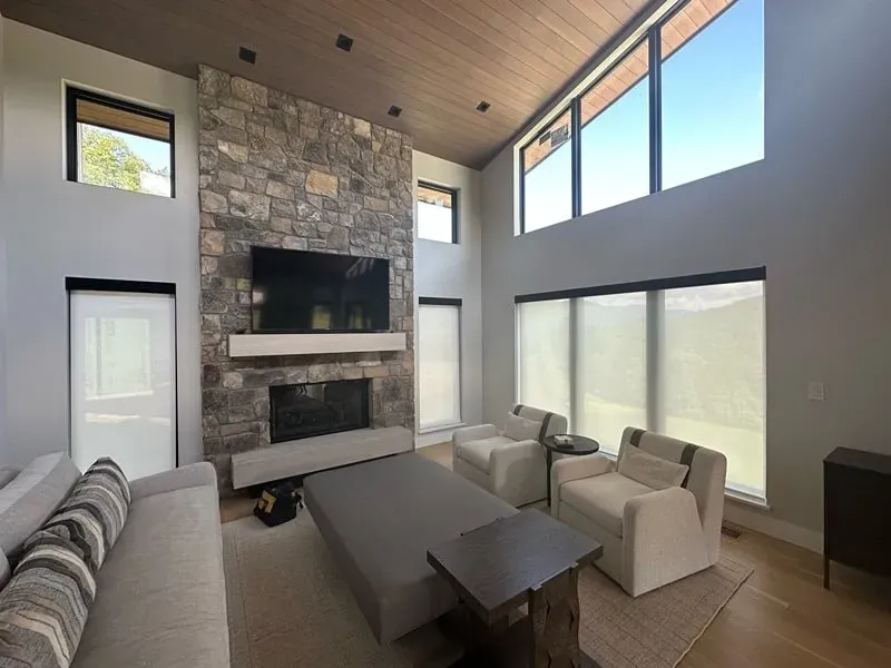 A modern living room with a stone fireplace, high vaulted wooden ceilings, large windows, and neutral-toned seating.