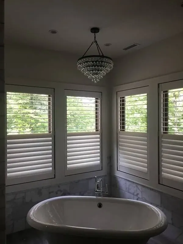 A standalone white bathtub sits below three windows with white shutters in a bathroom with a crystal chandelier.
