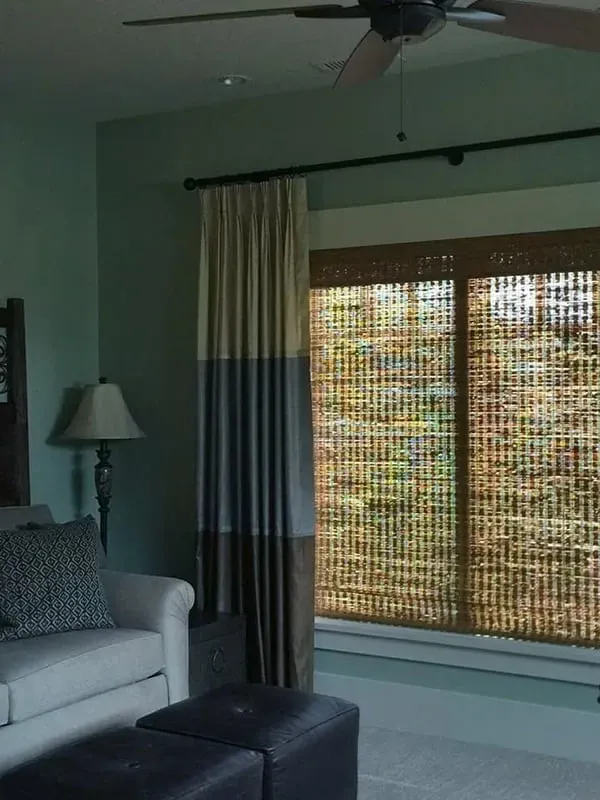 A living room corner with a neutral sofa, patterned pillow, black ottomans, table lamp, and window with tiered curtains.