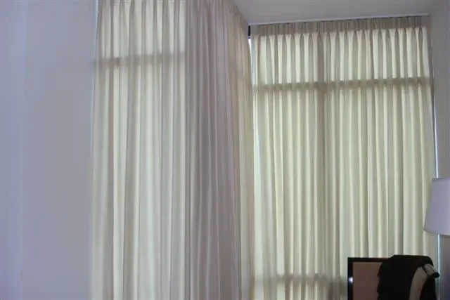 Floor-to-ceiling light beige curtains covering a corner window with a glimpse of a lamp and chair in the room.
