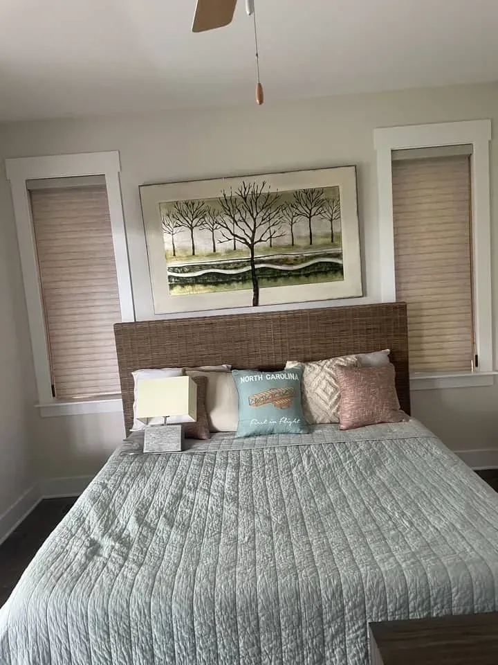 A bed with a wicker headboard, light blue quilted coverlet, and assorted pillows beneath a framed landscape wall art.