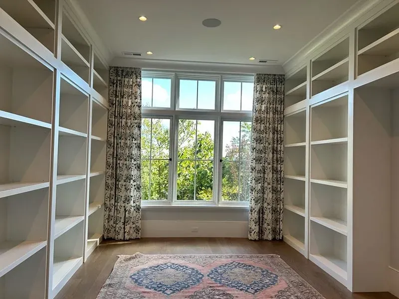 A bright home library with built-in white bookshelves on both sides, a large window with patterned curtains, and a rug.