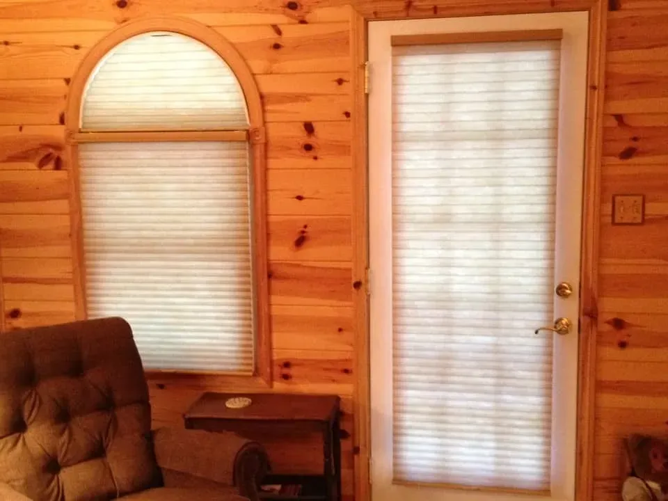 A view of a wood-paneled room with a tan armchair, a small side table, an arched window, and a door with light-filtering shades.