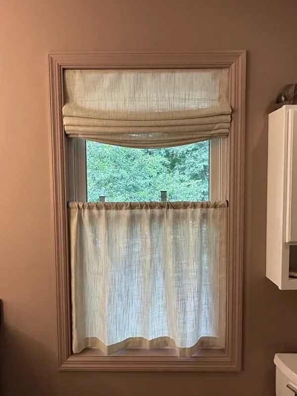 A window with a tan roman shade covering the top half and a gathered cafe-style sheer curtain covering the bottom half.