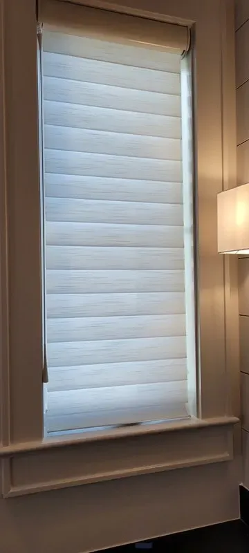 A white, horizontally pleated roller shade covering a window framed in white trim, with part of a wall lamp visible.