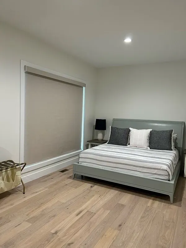 A grey bed with striped bedding sits on light wood floors in a room with a large window and a small bedside lamp.