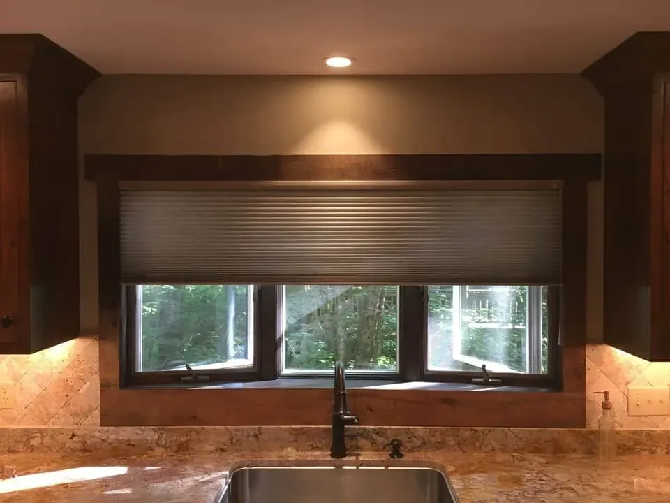 A dark wood-framed kitchen window above a sink, featuring a partially lowered pleated shade and tiled backsplash.