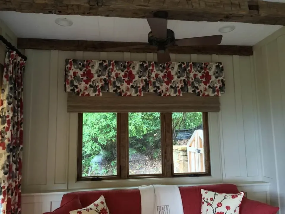 A red sofa sits beneath a window framed by floral patterned curtains and a matching valance, set against white walls.