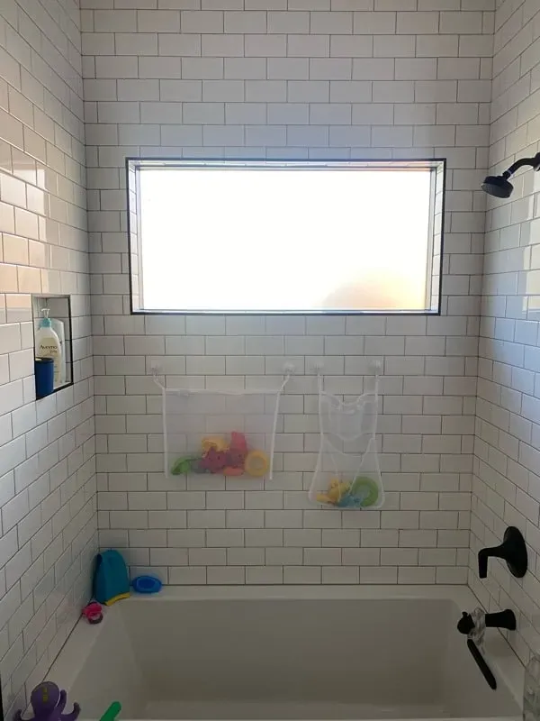 A white subway-tiled bathtub alcove with a window, two mesh toy storage bags, a recessed niche, and a shower faucet.