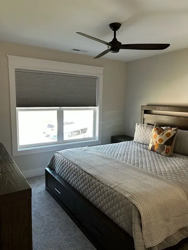 A bedroom with a queen bed featuring a grey quilted spread, a dark wood frame, a window with grey shades, and a ceiling fan.