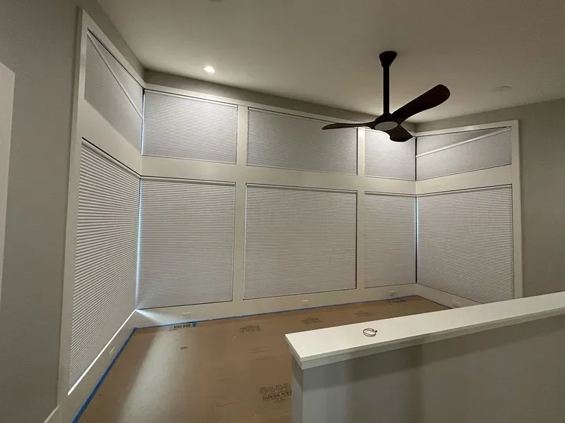 A corner wall of a room with large windows covered by grey horizontal blinds, a dark ceiling fan, and a low white wall.