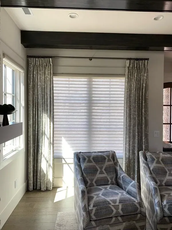 A window with sheer blinds and decorative curtains, flanked by two patterned armchairs in a room with dark ceiling beams.