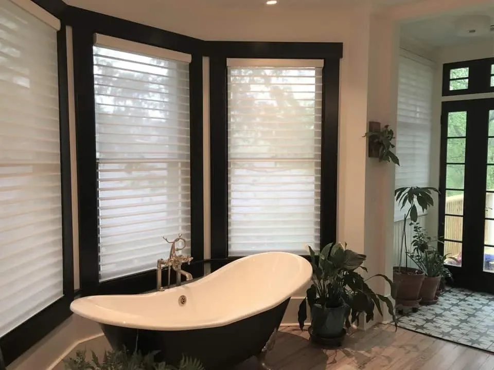 A black-and-white clawfoot bathtub sits by a window with white blinds and black trim in a sunlit bathroom with plants.