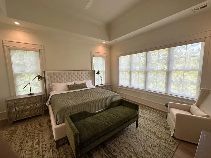 A bright bedroom with a tufted bed, olive green bedding and bench, a patterned rug, and large windows with white shades.