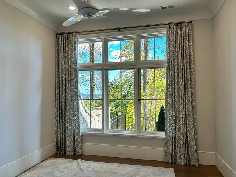A window with patterned drapes overlooks a view of trees under a blue sky, set in a room with white walls and flooring.